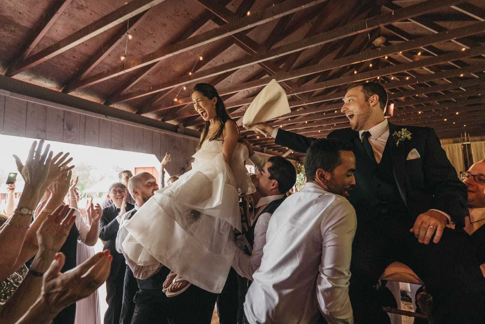 Bride and groom are lifted up on chairs during hora at wedding reception, groom looking excited and bride smiling widely but nervously.