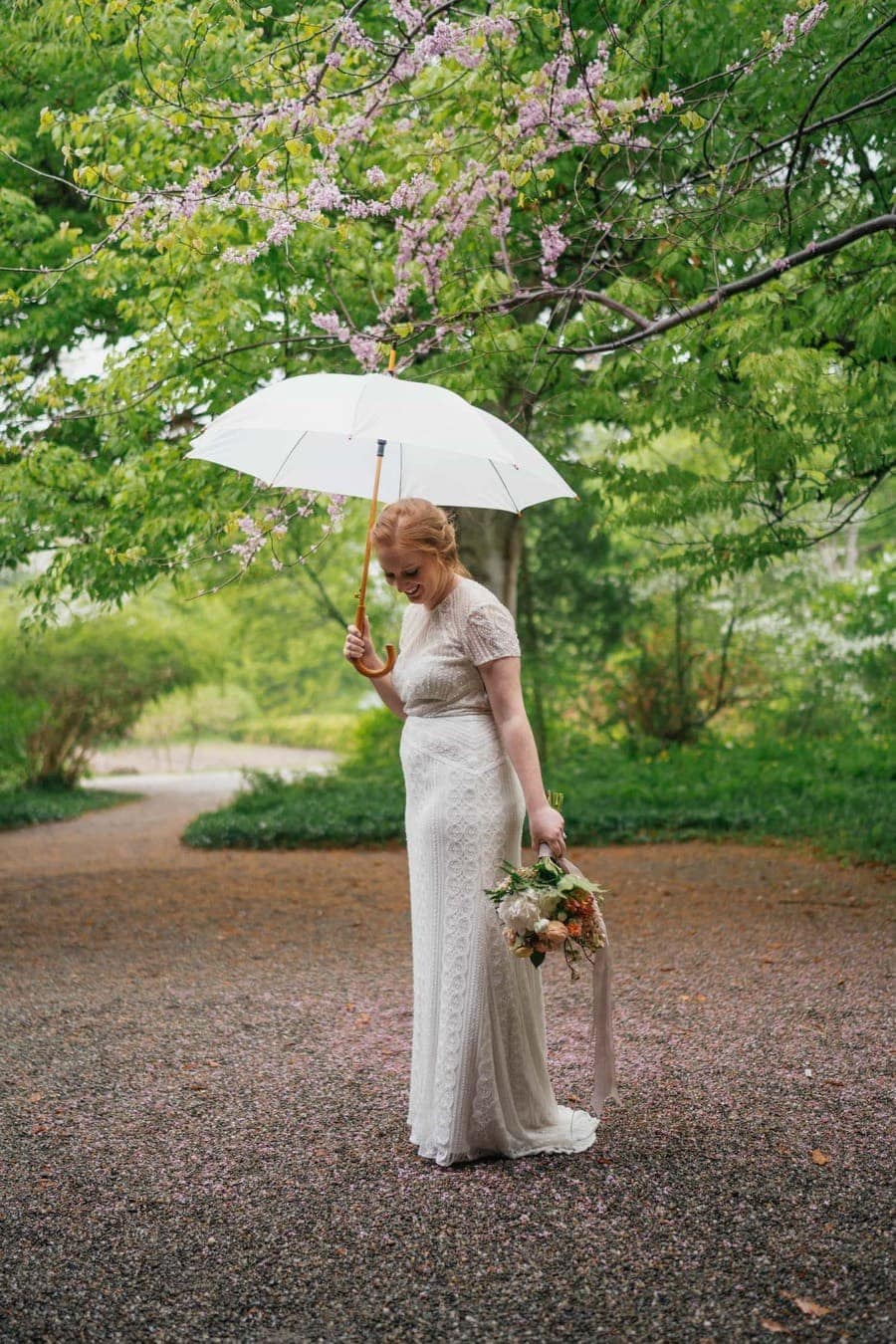 Bride in wedding dress smiles and looks at ground, holding bouquet and white umbrella.