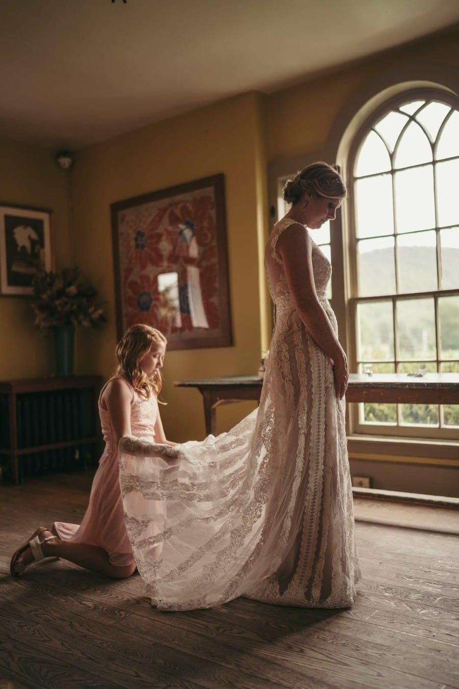 Flower girl fixes bride's dress as light from window comes into room and shines through lace dress before wedding ceremony.
