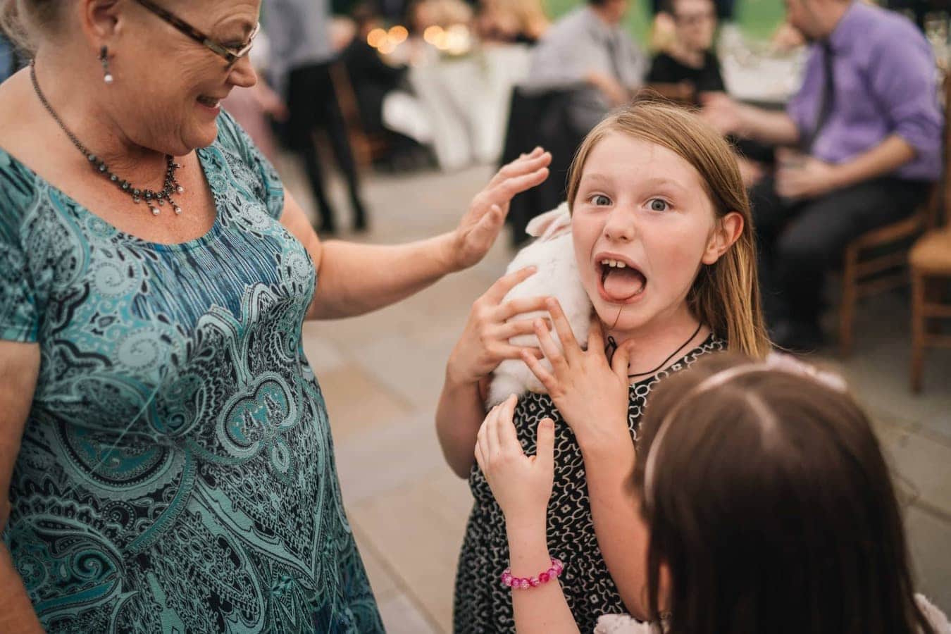 Little girl holding a white rabbit on her shoulder makes funny face by sticking out tongue during wedding reception in Hudson Valley.