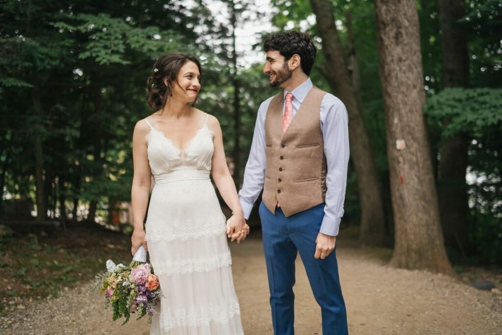Bride and groom hold hands and smile at each other on forest path.