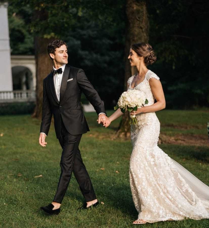 Bride and groom hold hands, groom looking back at bride, walking past A Private Estate's house in Hudson Valley.