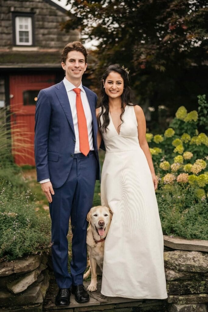 Bride and groom stand together and smile with dog outside of Catskill wedding venue.