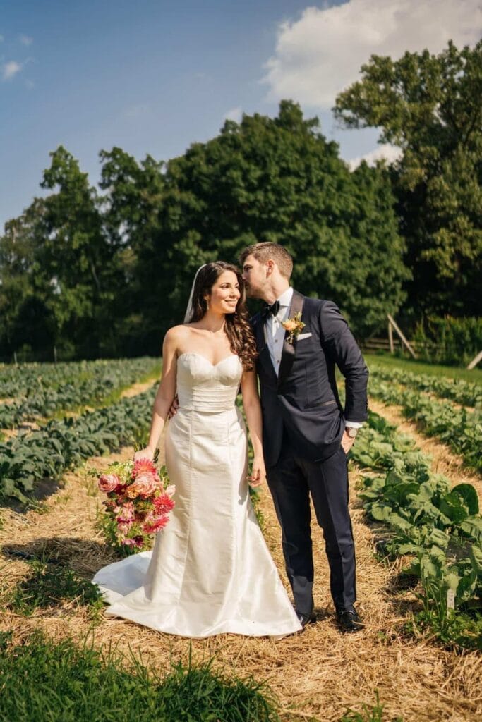 Groom whispers to bride and bride smiles as they stand together in farm fields of Hudson River wedding venue.