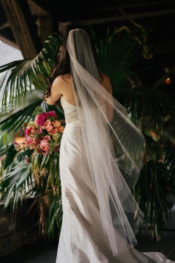 Bride in dress and long dress turns back to camera while standing in palm plant.