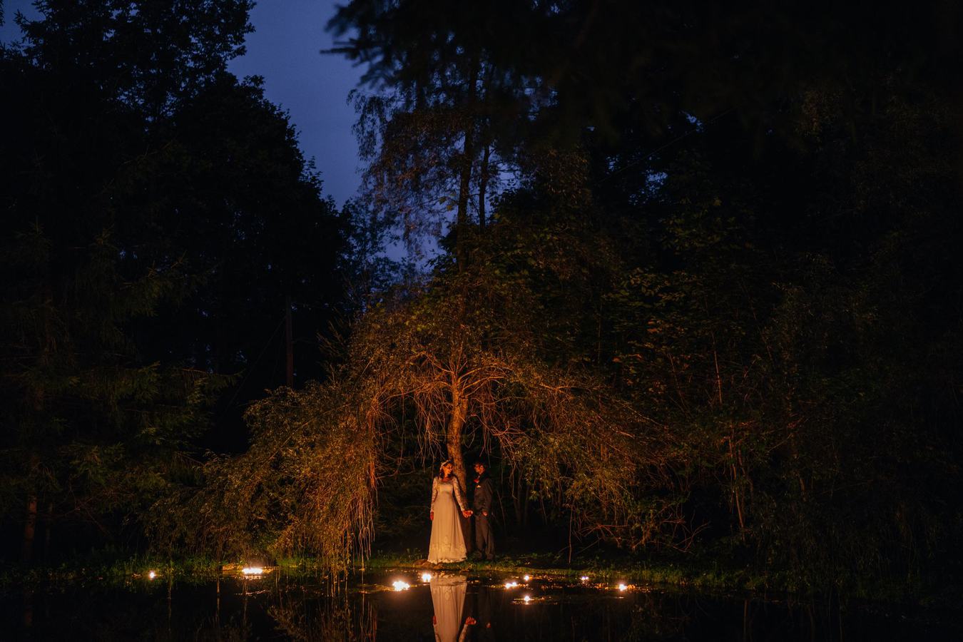 Bride and groom stand underneath tree by candle lit pond and hold hands at night at Catskill wedding venue.