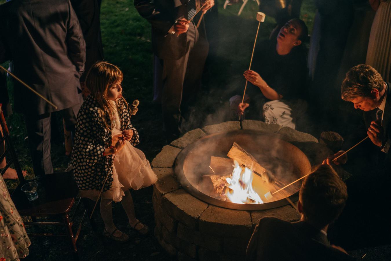 Wedding guests sit around fire pit to make s'mores, and a little girl blows on her marshmallow to cool it down after cooking it.
