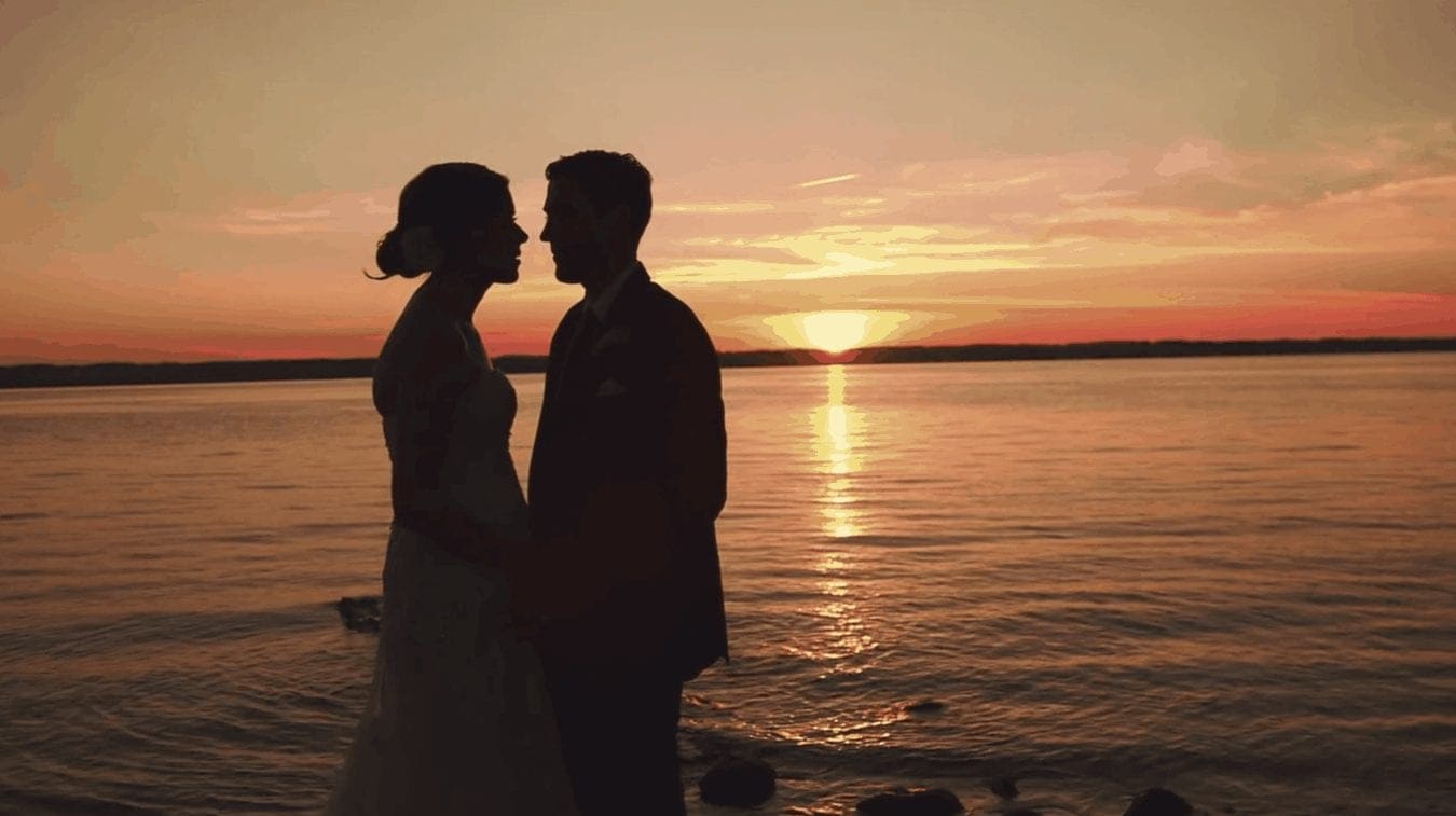 Bride and groom silhouette against sunset on lake.