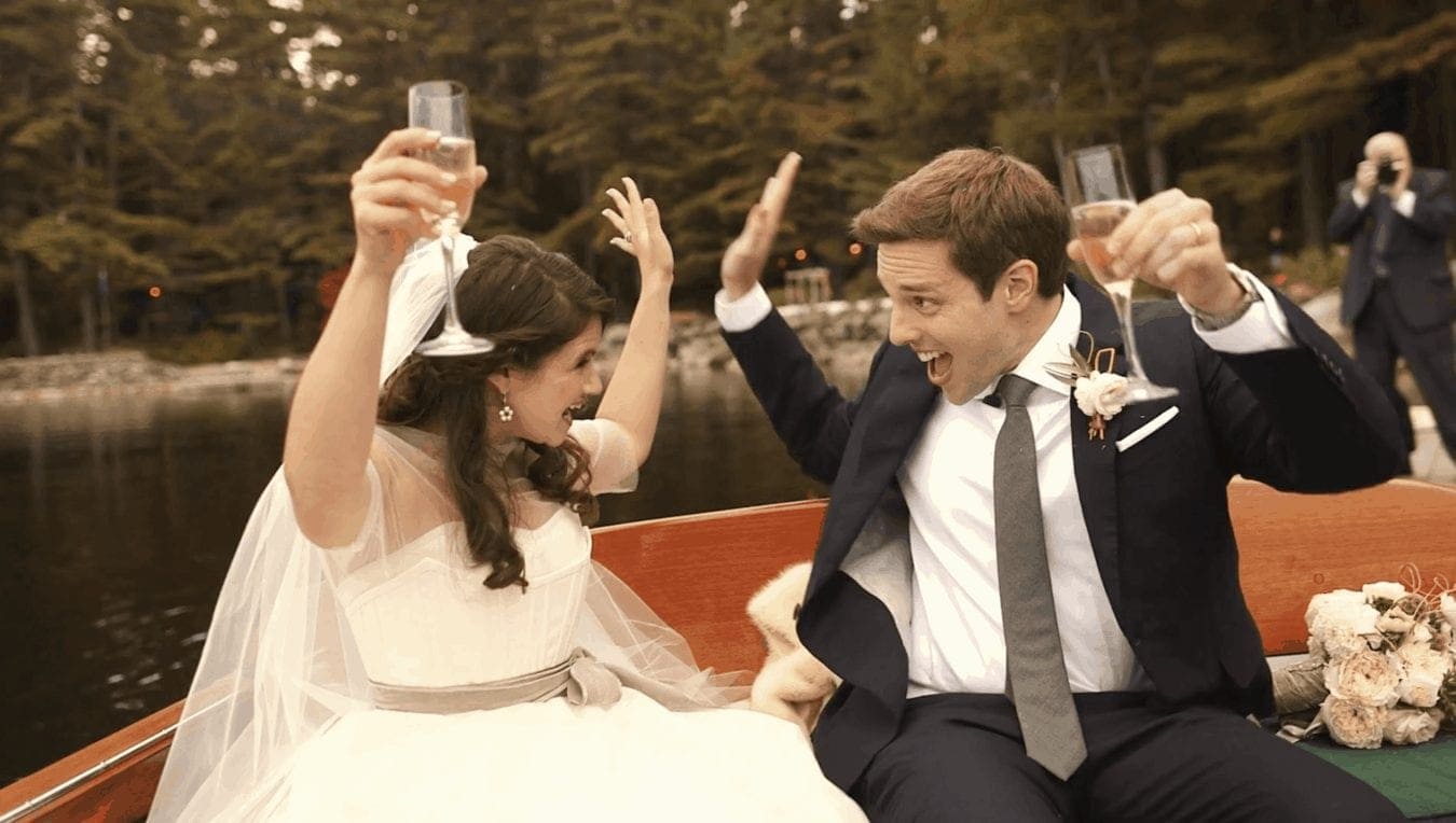 Bride and groom in boat smile at each other and throw their arms up in celebration.