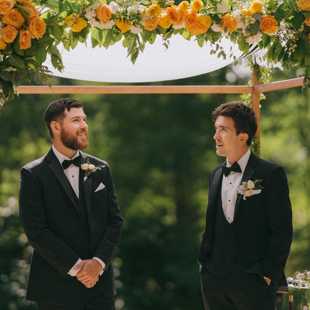 Groom and friend smiling under floral chuppah at wedding.