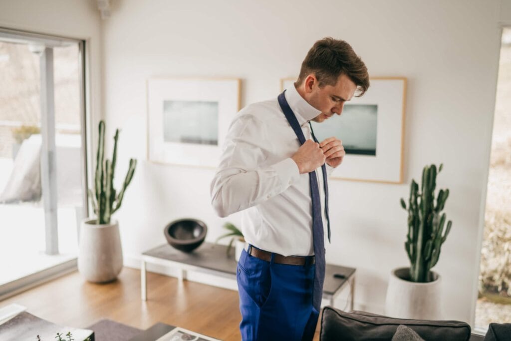 Groom begins putting on tie before wedding ceremony in mid-century home in Hudson Valley.