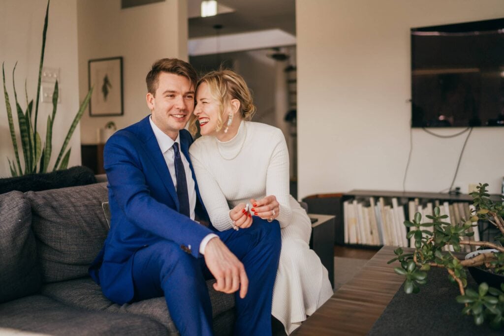 Bride smiles and laughs at groom, and groom smiles, both sitting on couch inside midcentury home in Hudson Valley.