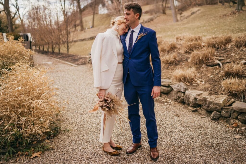 Bride rests smiles and rests head on grooms should, both standing on gravel walkway in Hudson Valley winter scenery.