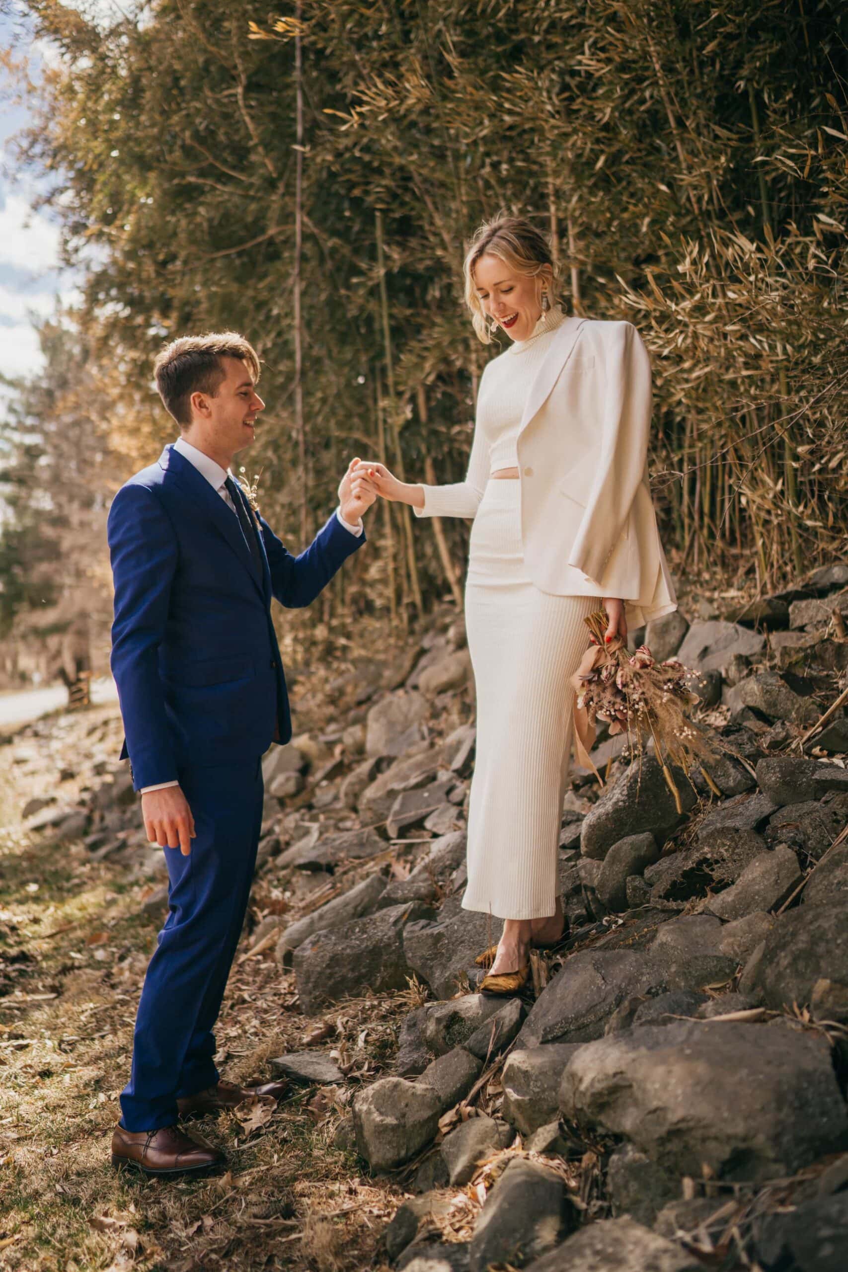 Bride stands on rocks and smiles, groom holds her hand as she walks down.