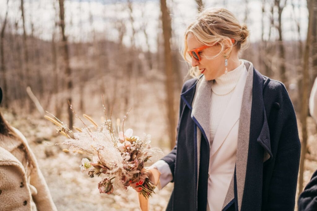 Bride in dress and orange sunglasses looks at and holds bouquet in Hudson Valley winter forest.