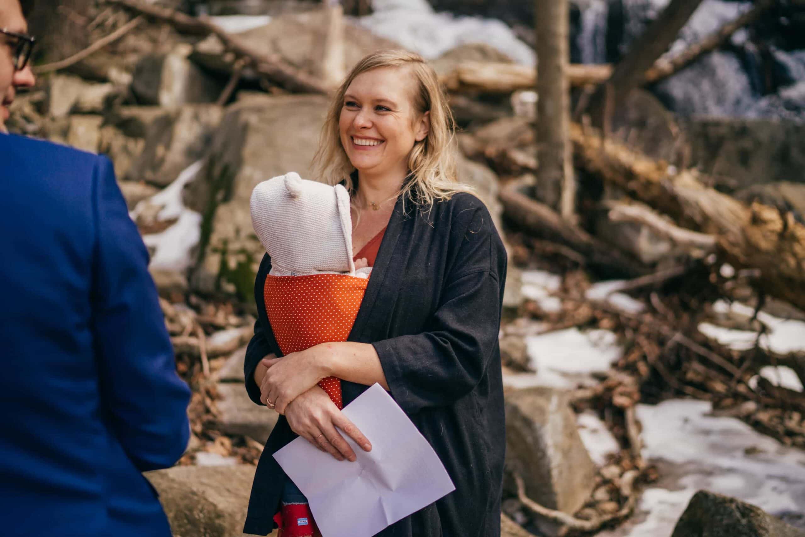 Wedding officiant with baby strapped to her smiles at wedding couple in winter Hudson Valley forest.