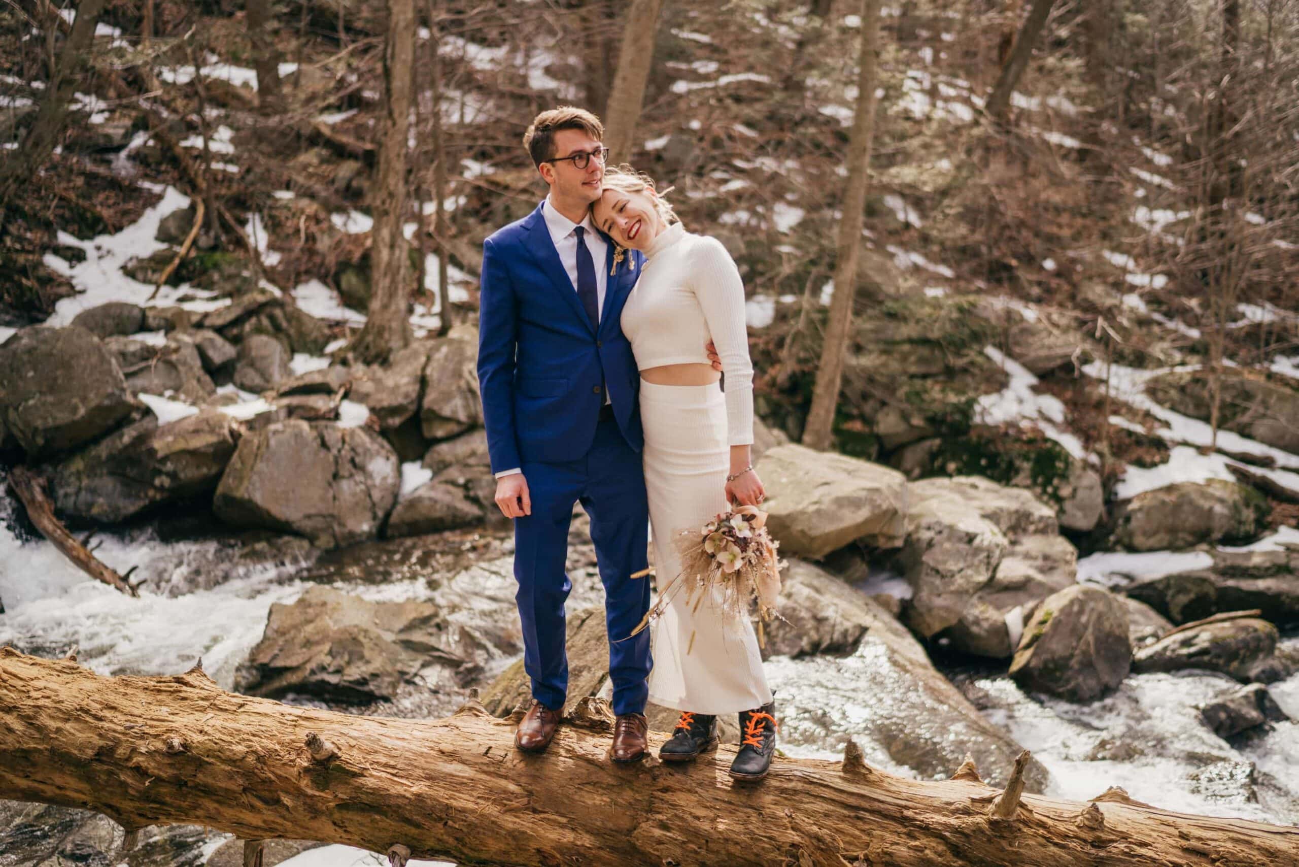 Bride in dress and hiking boots smiles and leans on groom's shoulder as he puts his arm around her waist, standing on a log over a river in winter Hudson Valley forest.