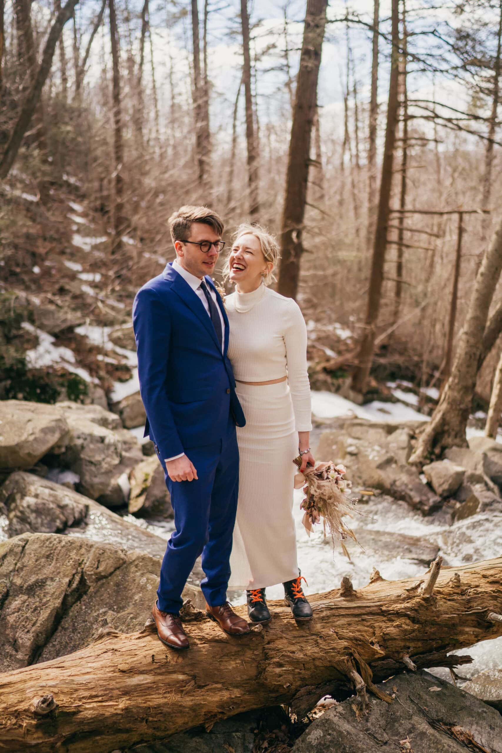 Bride in dress laughs at groom in blue suit as they stand on a log over a river in winter Hudson Valley forest.