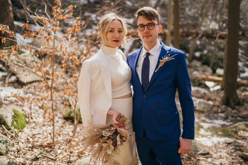 Bride in white dress and groom in blue suit look seriously at the camera and stand together in winter Hudson Valley forest.
