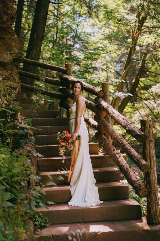 A bride ascends a rustic staircase, curving around a rock. She looks back toward the camera, holding a colorful bouquet in tones of pink and orange. In the background, we see lush summer woods.