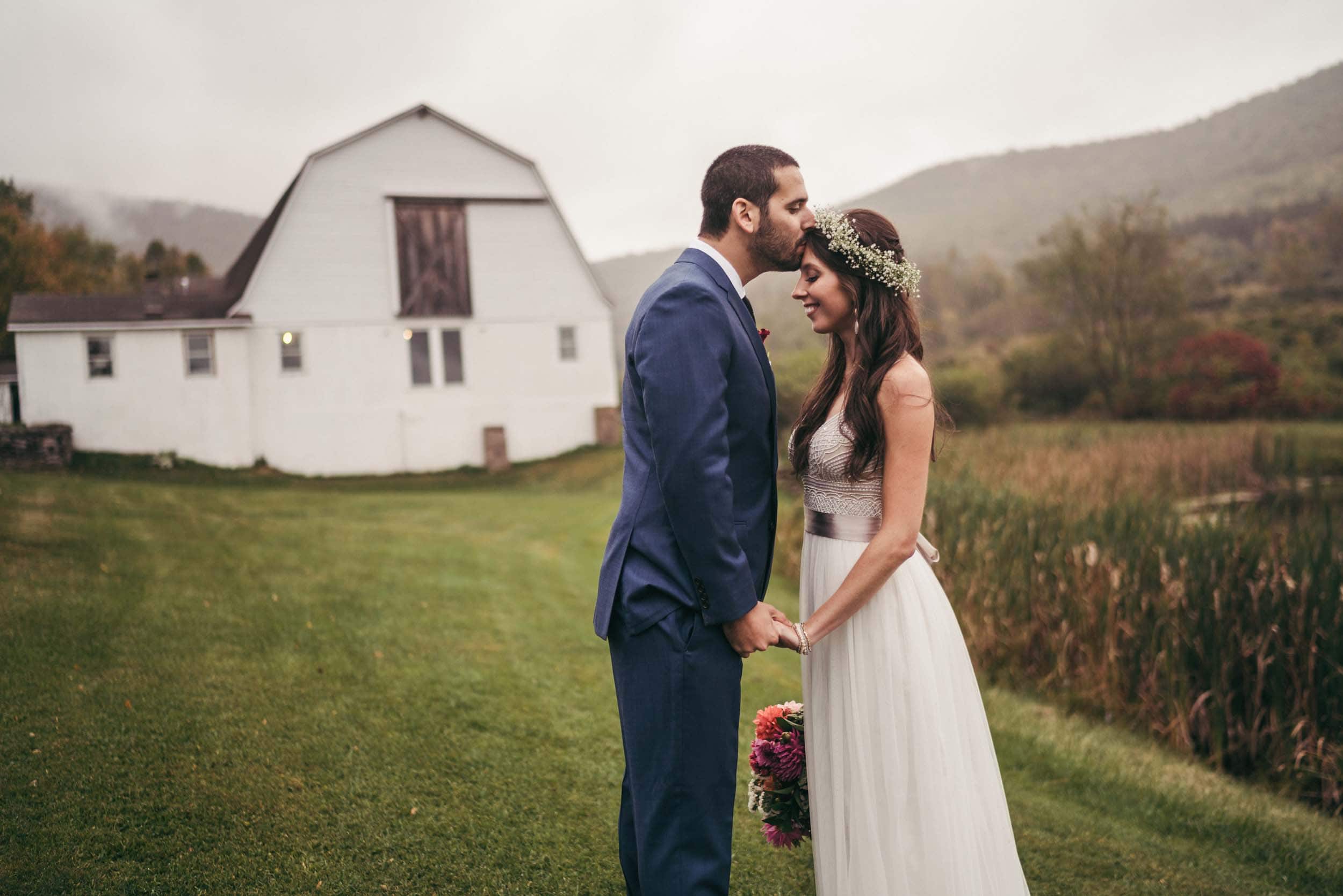 A bride with long brown hear wearing a flower crown of baby's breath and a sleeveless white wedding dress receives a forehead kiss from a groom in a blue suit. Behind them is a barn, along with misty foothills and farmland.