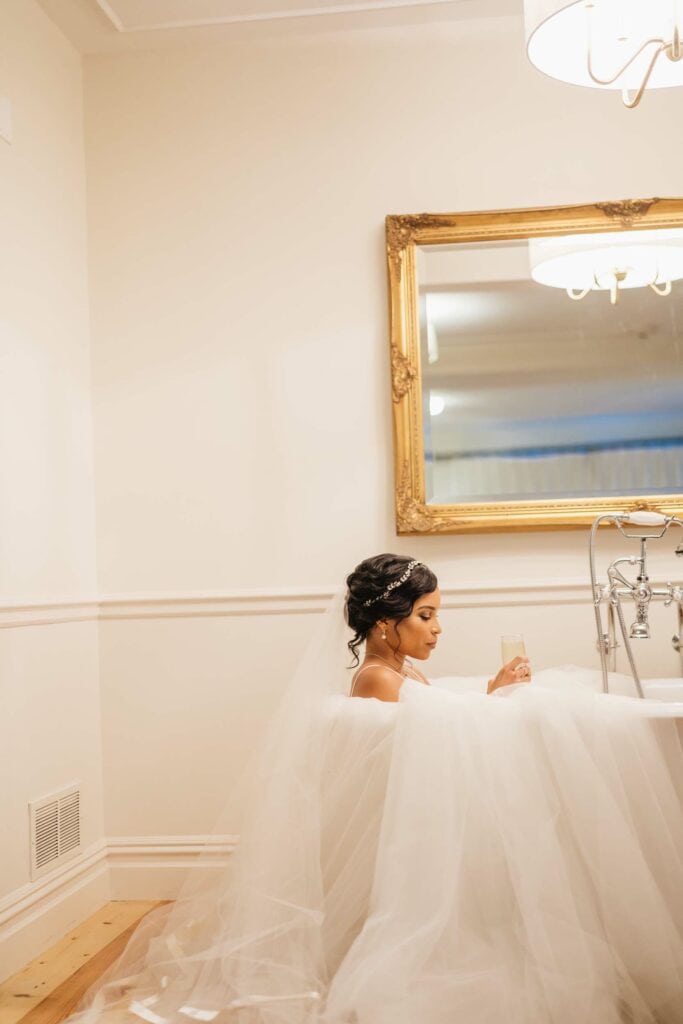 A bride swathed in tulle enjoys a quiet moment sitting in an old-fashioned (dry) claw-foot bathtub while drinking a glass of champagne. Her veil trails over the back of the tub behind her.
