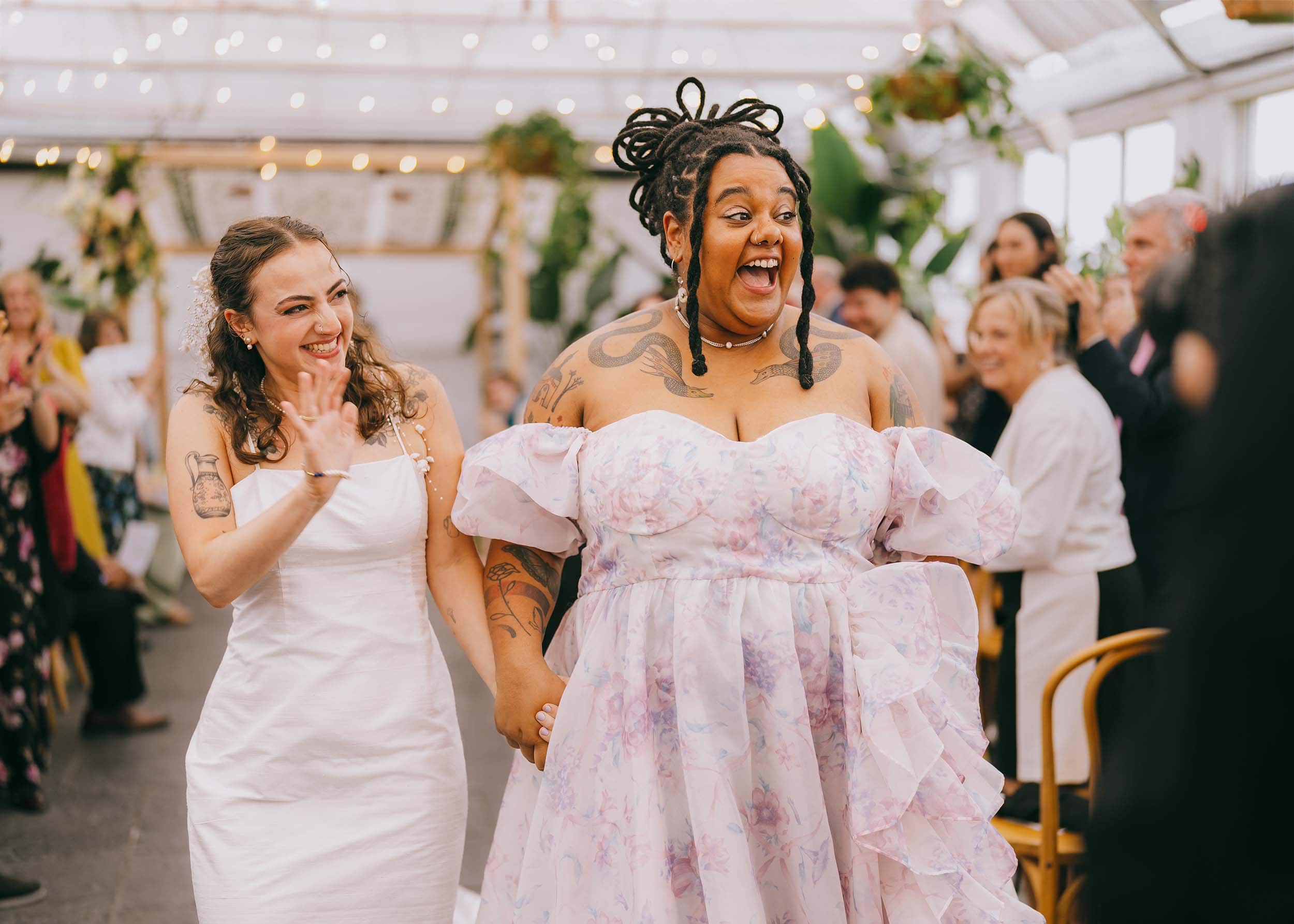 Eve and Cece beam with joy as they stroll down the aisle, greeted by standing guests who warmly congratulate them on their newly sealed union.