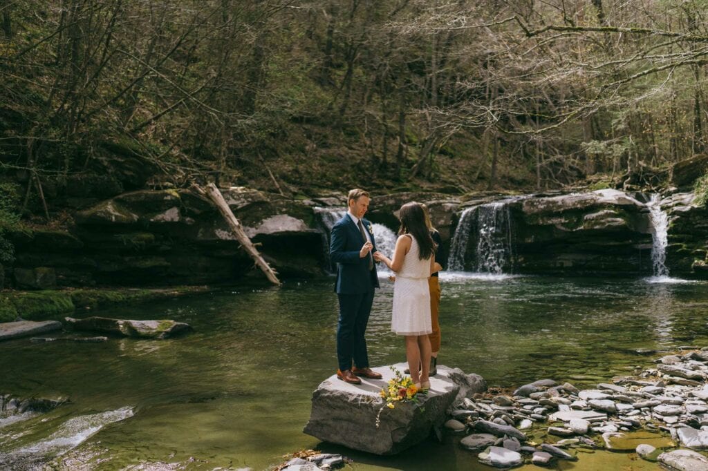 A couple elopes to the Hudson Valley. They exchange vows on a boulder at the edge of a pool. Behind and beside them, small waterfalls cascade. 