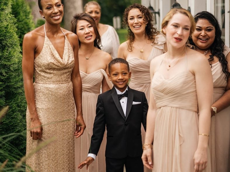 Bridesmaids and nephew of bride admire bride as she shows them her wedding dress.