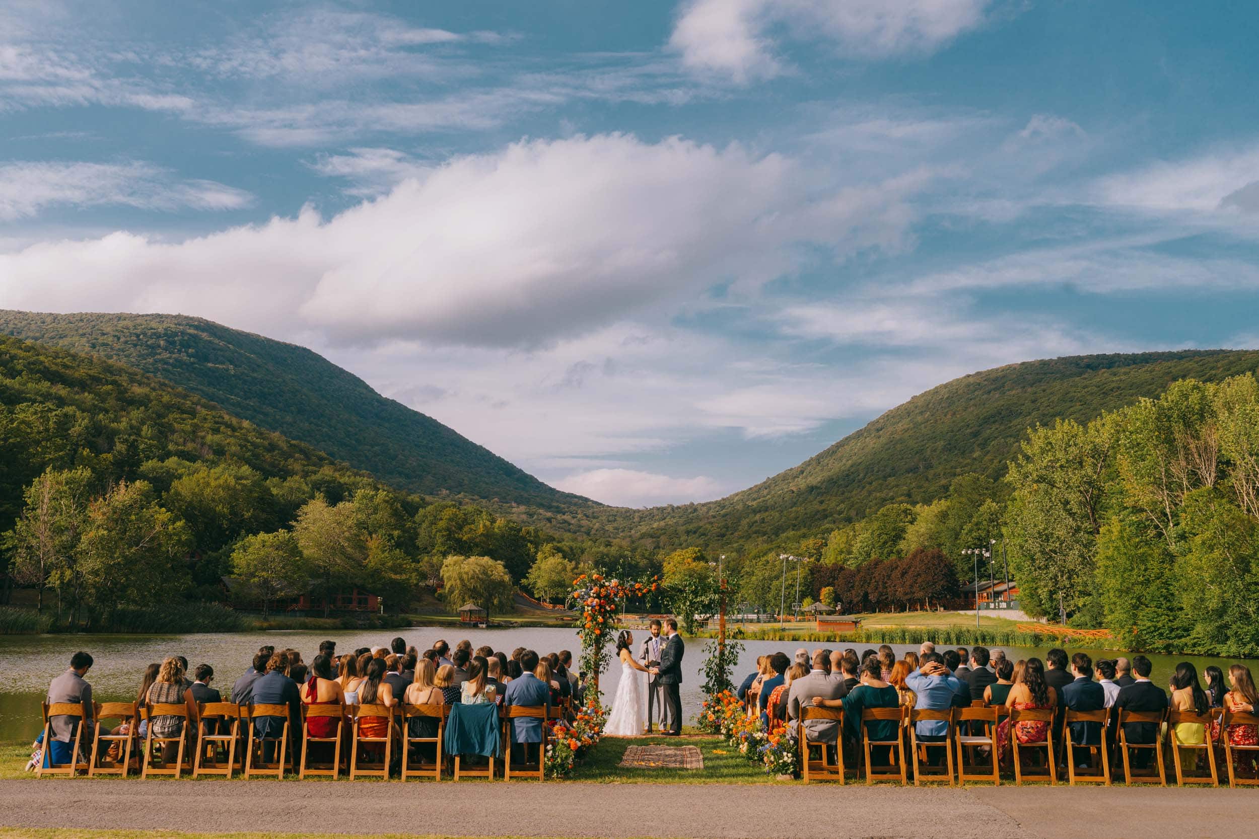 A couple stands under a flower arch at their lakeside ceremony, surrounded by green hills and family.