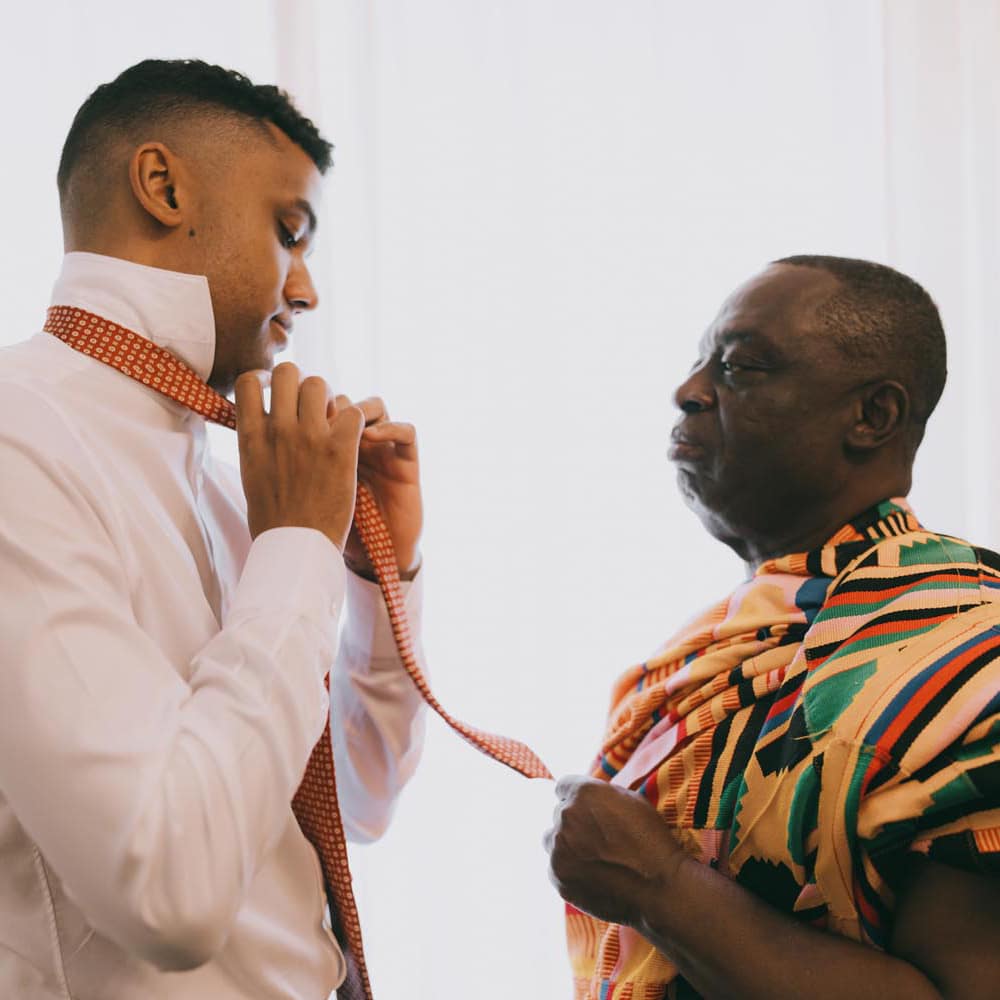 Father of the groom in traditional attire helps his son with his tie.