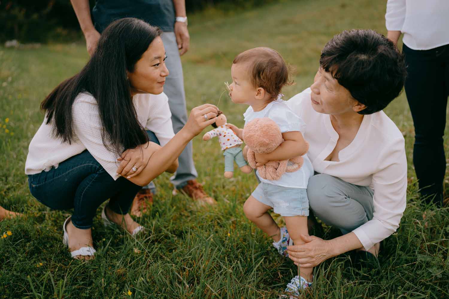 A family member, couched down to kid level, holds a flower up to a child's nose so that they may smell it, while another family member holds the child.