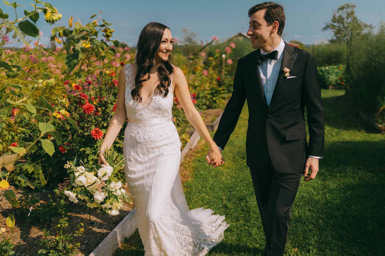 A bride and groom walk hand in hand through a flower garden under a blue cloudless sky. 