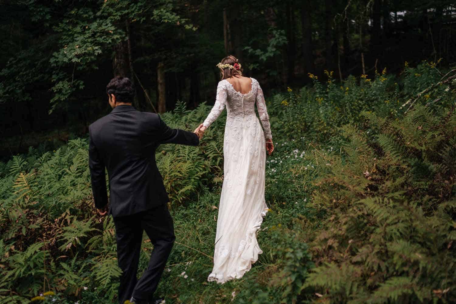 Action: View more details about elopements. Image: A bride in a long lace dress with a short train leads her groom up a slope into the woods by the hand, surrounded by woodland edge plants, with trees ahead.